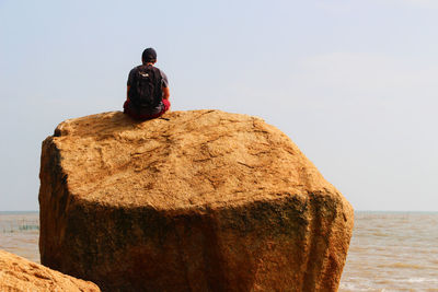 Rear view of backpacker sitting on rock against clear sky