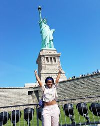 Low angle portrait of smiling woman standing with arms raised against statue of liberty