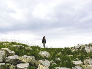 Mid distance view of young woman standing on grassy field against cloudy sky