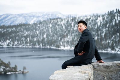 Portrait of young man sitting on retaining wall over lake