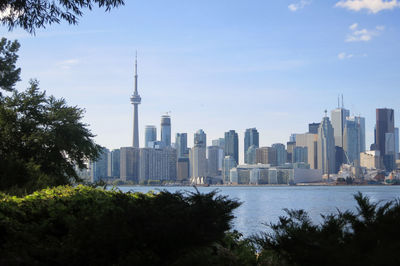 Cn tower amidst buildings in front of river and trees against sky on sunny day