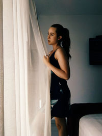 Side view of young woman holding curtain in bedroom
