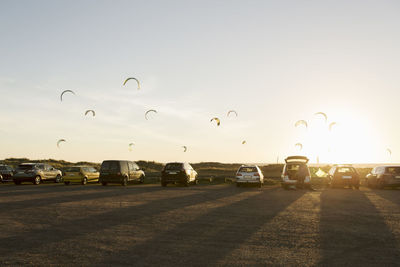 Cars parked on beach against parachutes in sky during sunset