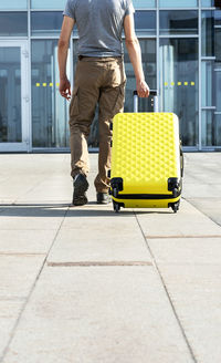 Man traveler in casual carrying yellow suitcase next to entrance to airport outside  vacation mockup