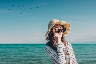 Young woman in hat and sunglasses standing against sea