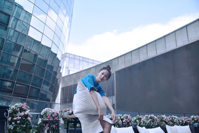 Portrait of young woman holding pillow while standing against modern buildings