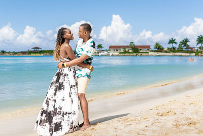 Portrait of a young smiling couple standing on the beach against the sea and clear sky