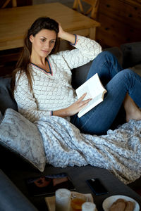 Woman with book looking away while sitting on sofa at home