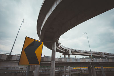 Low angle view of road sign below bridges against cloudy sky