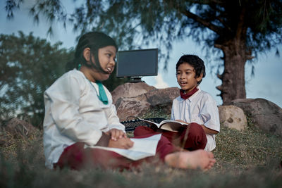 Happy friends studying while sitting by tree outdoors