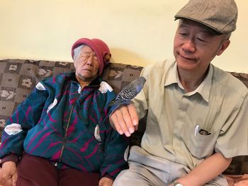 Senior man with budgerigar sitting by woman on sofa at home