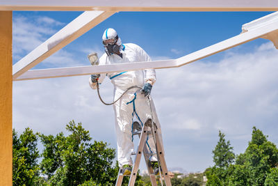 Anonymous employee in protective uniform and mask standing on ladder and using air brush to paint frame structure against cloudy sky on sunny day