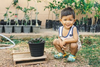 Portrait of cute smiling baby boy crouching by potted plant on field