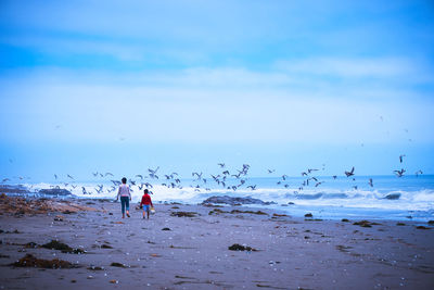 Rear view of mother and son walking while birds flying over beach against sky