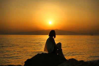 Silhouette woman sitting on shore against orange sunset sky