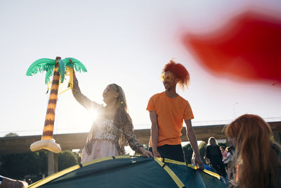 Woman holding balloon while making tent with friends against sky during sunny day