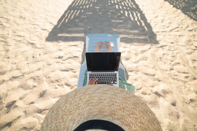 Top view of slender young caucasian woman in hat works on his laptop resting on sandy beach 