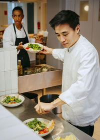 Male chef passing salad bowl to female chef while finishing salad at commercial kitchen