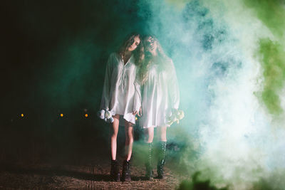 Full length portrait of female friends standing amidst smoke on land at night