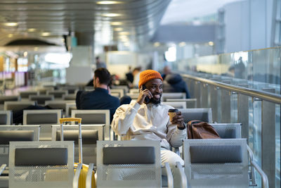 Smiling black traveler man drinking coffee waiting for flying at airport talking on mobile phone