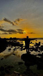 Rear view of man standing at beach against sky during sunset