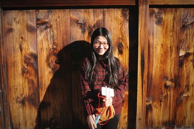 Portrait of smiling woman holding monopod and mobile phone while standing against wooden wall