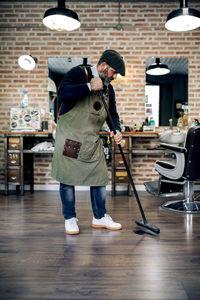 Full length of middle aged bearded hispanic male barber in apron and beret sweeping hair from floor with broom in traditional barbershop