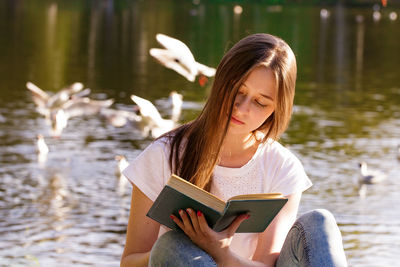 A young woman sitting by the lake reading a book