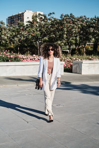 Full body hispanic female manager in trendy white suit and sunglasses with curly hair looking away while walking on the street on sunny day in city