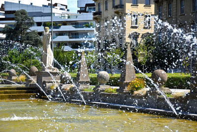 Fountain in park by lake against buildings