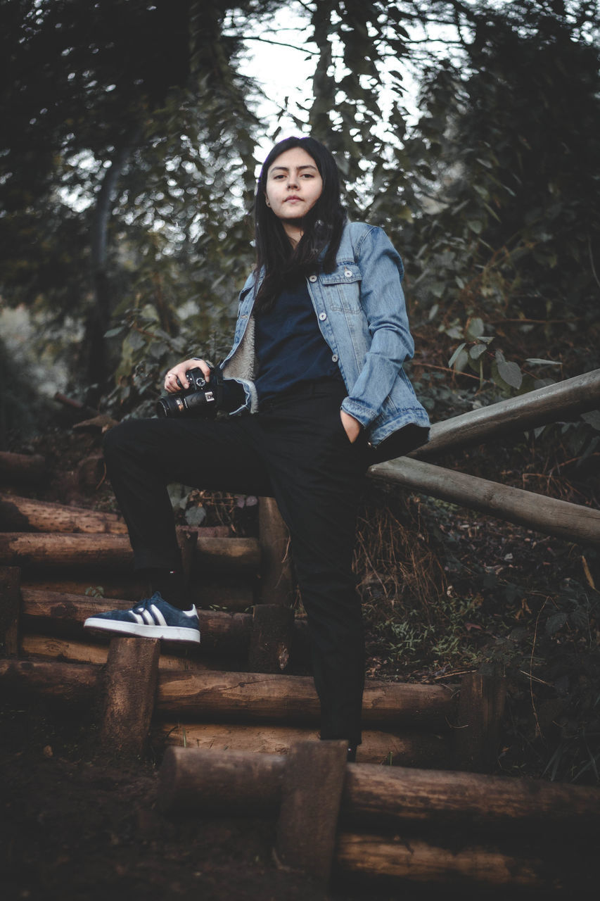 full length, one person, young adult, casual clothing, tree, real people, front view, plant, leisure activity, portrait, lifestyles, forest, nature, looking at camera, sitting, day, land, young women, clothing, outdoors, contemplation, hairstyle