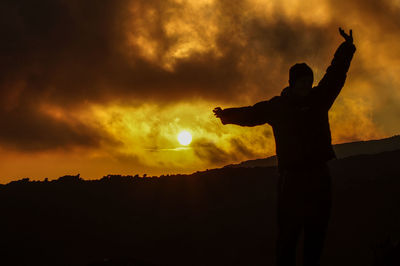 Silhouette man with arms outstretched standing against orange sky