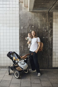 Smiling man with hand in pocket carrying daughter while standing by stroller in front of wall
