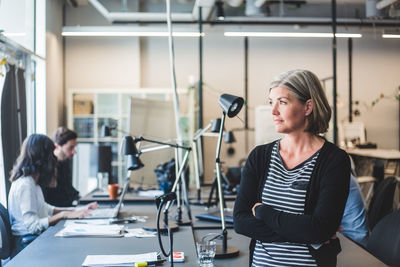 Confident businesswoman looking away while standing against desk in creative office