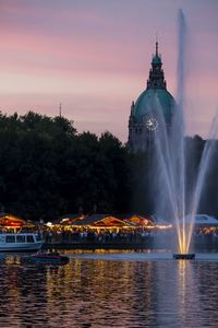 Illuminated building by river against sky during sunset