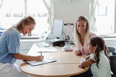 Smiling pediatrician writing prescription while girl sitting with mother at desk in clinic