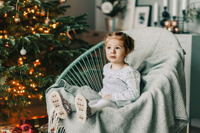 A little girl is sitting in an armchair and smiling against the background of a christmas tree 