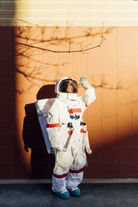 Female astronaut in space suit shielding eyes from sunlight while standing by wall