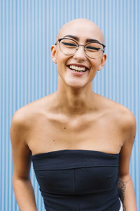 Cheerful young woman with shaved head wearing eyeglasses in front of blue wall