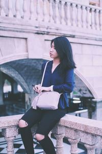 Thoughtful young woman looking down while sitting on retaining wall in city