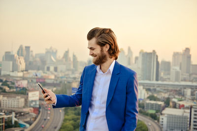 Businessman using smart phone while standing against cityscape during sunset