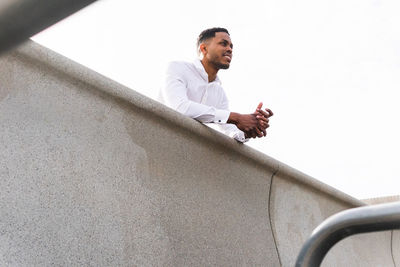 From below of confident african american male entrepreneur in elegant clothes looking into distance while standing near contemporary high rise building on sunny street