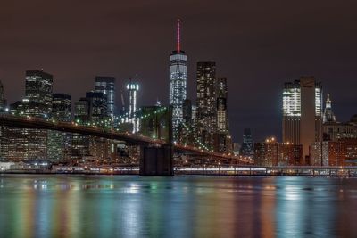 Brooklyn bridge over river against illuminated one world trade center in city