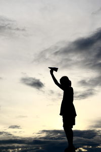 Silhouette girl holding paper airplane against sky