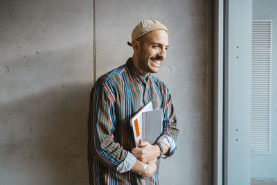 Cheerful male adult student holding book standing in front of gray wall at college