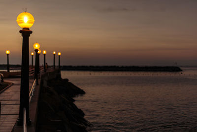 Illuminated street light on pier by sea against sky during sunset
