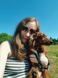 Portrait of young woman embracing puppy while sitting on grassy field against clear blue sky