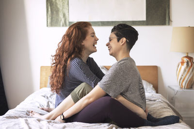 Side view of cheerful lesbians looking at each other while sitting on bed