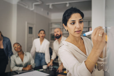 Female entrepreneur writing on whiteboard during meeting with coworkers in office