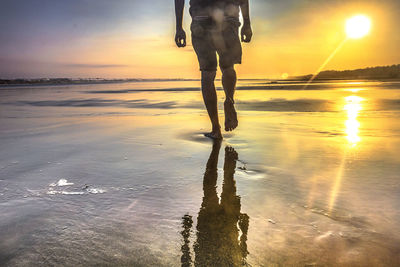 Low section of man walking on shore against sunset sky